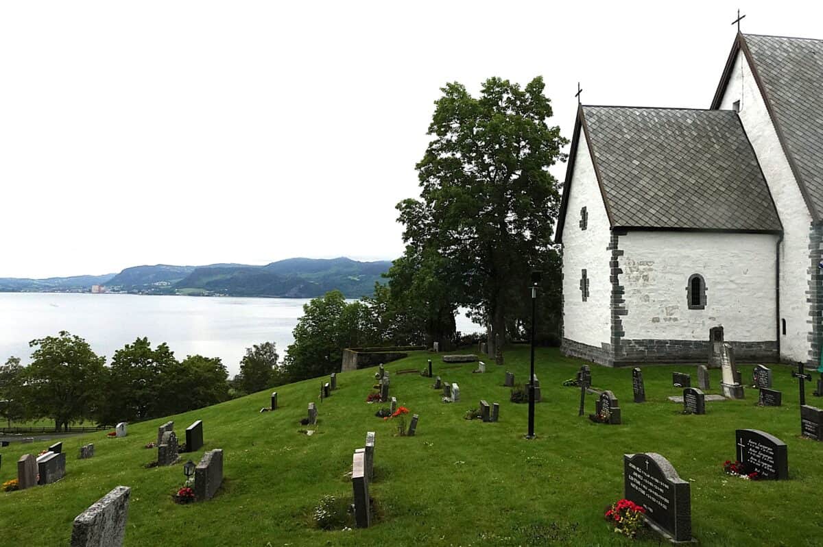 Byneset church is a medieval white, plastered stone church, built 1140-1180, with a slate roof, overlooking the Trondheimsfjord in Trøndelag, Norway. Image showing the graveyard and the view to the fjord and the modern industral landscape.